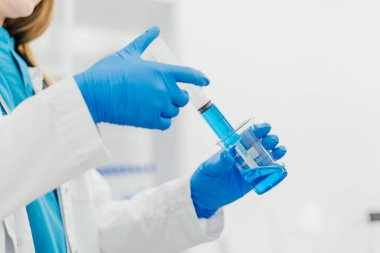 Mavi eldiven giyen, şırınga tutan ve mavi sıvıyla dolu bir laboratuvar şişesi olan ellerin yakın çekimi. Görüntü, tıbbi ve laboratuvar prosedürlerindeki hassasiyet ve profesyonelliği gösteriyor. Yüksek