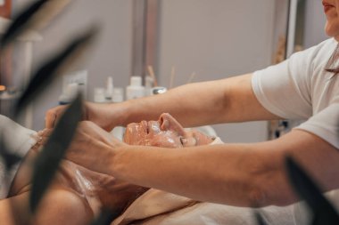 Yüz bakımı içeren sakin bir spa sahnesi. Bir terapist nazikçe bir maske takar müşterinin yüzüne, kaplıca ürünleri ve rahatlatıcı bir atmosfer ile çevrili..