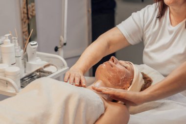 Kaplıcada yüz bakımı gören bir kadın. Bir güzellik uzmanı, cilt ürünleriyle çevrili tedavi masasında dinlenirken yüzüne maske takar..