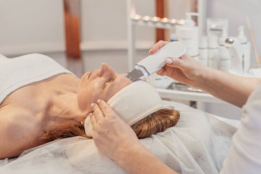 Kaplıcada yüz bakımı gören bir kadın. Bir terapist, cilt bakımı ürünleriyle çevrili bir tedavi masasında dinlenirken, yüzünde bir el bilgisayarı kullanır..