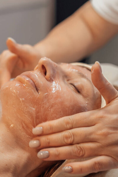 A woman receiving a facial treatment at a spa, with a focus on relaxation and skincare. The aestheticians hands are gently applying a gel or mask to the womans face, highlighting a serene atmosphere