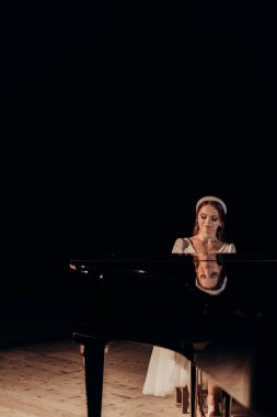 Beyaz elbiseli bir piyanist zarif bir şekilde kuyruklu bir piyanonun başında oturuyor, onun sakin ifadesi enstrümanın parlak yüzeyine yansıyor. Spot ışıkları zamansız bir ortam yaratır. Akan bir piyanist