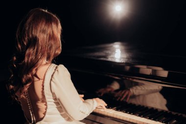 Zarif beyaz bir elbise içinde inci detaylarıyla bir piyanistin arka görüntüsü. Elleri spot ışıkları altında piyano çalıyor, yansıtıcı ve huzurlu bir atmosfer yaratıyor.
