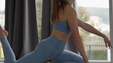 Kapalı mekanda yoga yapan, güvercin pozisyonunda gerinen bir kadın. Açık mavi bir spor kıyafeti giyiyor, esneklik ve denge odaklı. Doğal ışık pencereden süzülür