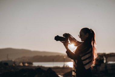 Bir kadın fotoğrafçı, gün batımının altın ışığı siluetini aydınlatırken kamerasını ayarlıyor. Açık havada, uzak tepelerden oluşan sakin bir zemin ve ışıldayan bir suyla duruyor. Uzun saçları ve...