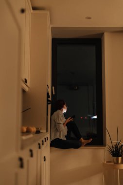 Bir kadın gece rahat bir mutfakta büyük bir pencerenin yanında oturuyor, elinde akıllı bir telefon var. Sevdiği biriyle internete bağlanırken yüzündeki o düşünceli ifadeyi vurguluyor. Bu huzurlu aşk anı