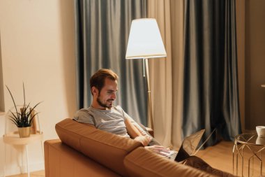 Bir adam dizüstü bilgisayarıyla koltukta rahatça oturur çalışırken ya da internette dolaşırken gülümser. Sıcak aydınlatma ve sakin atmosfer, üretkenlik ve bağlantının rahat bir akşamını vurguluyor