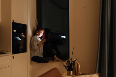 Bir kadın karanlık bir mutfağın pencere eşiğinde oturuyor, yüzü akıllı bir telefonun parıltısıyla aydınlanıyor. Uzaktan sevdikleriyle huzurlu bir bağ anı.