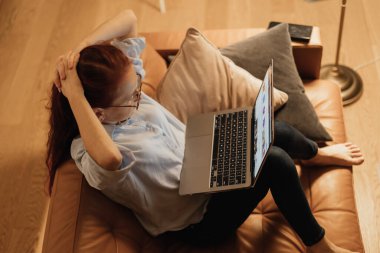 Uzun saçlı bir kadın deri koltukta oturup bilgisayarını karıştırıyor. Sıcak aydınlatma ve rahat yastıklar rahat bir atmosfer yaratıyor. Çevrimiçi veya romantik anları keşfetmek için mükemmel..