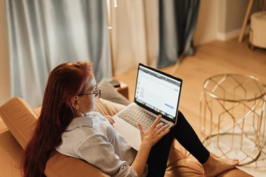 Uzun saçlı bir kadın deri bir kanepede yatarken internetteki bağlantıları keşfediyor. Sıcak ışıklandırması ve dizüstü bilgisayarı olan rahat bir ev düzeni sanal ilişkiler ve uzun süreli ilişkiler için mükemmel bir ortam yaratır.