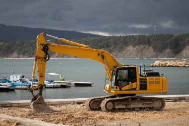 Canlı bir sarı kazıcı, gelecek için altyapı ve kıyı kalkınma projelerine sağlam bir yatırımı simgeleyen pitoresk bir kıyı şeridinin yanında duruyor.
