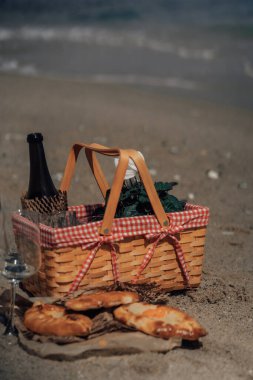 Kumlu bir plajda kareli bez, şarap şişesi ve hamur işleriyle dolu bir piknik sepeti. Kıyı pikniği, dokuma sepetli şarap kadehleri ve khachapuri kumlu sahilde arka planda deniz olan kağıt seti.