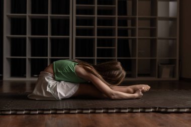 Yeşil bluzlu ve beyaz şortlu genç kız loş ışıklı spor salonunda derin esneme egzersizi yapıyor meditasyon havasını yakalıyor ve karanlık jimnastik salonunda sakin dengeyi sağlıyor.