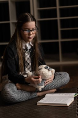 Okul üniformalı genç bir kız, elinde pelüş bir oyuncakla yerde bağdaş kurarak oturur. Okuma, erken eğitim, duygusal gelişim ve kapsayıcı öğrenme hakkında bilgi verir.