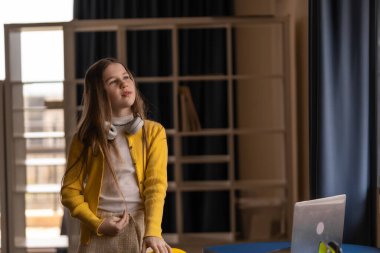 Genç bir öğrenci dizüstü bilgisayarı ve kulaklığı olan bir masanın başında duruyor. Eğitimsel yolunu ve modern bir sınıfta doğal ışık ile gelecek olasılıklarını yansıtıyor.