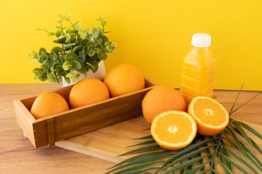 Bir şişe portakal suyu, yeşil tropikal yapraklı ahşap bir yüzeye yarı yarıya ve bütün portakalların yanına yerleştirilir. Canlı sarı arka plan, bölgeye enerjik, taze ve sağlığa odaklı bir his katıyor.