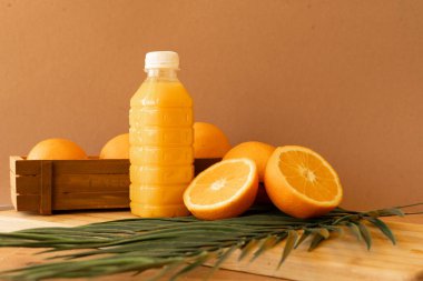 Taze portakal suyuyla dolu şeffaf bir plastik şişe ahşap bir yüzeyde duruyor ve yeşil palmiye yapraklarıyla tamamlanmış portakallar narenciye çiçeğini kutluyor.