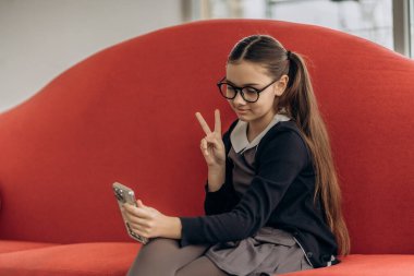Uzun saçlı, gözlüklü, rahat iç mekanında çekilmiş akıllı telefon fotoğrafı için barış jesti pozu veren genç ve şık bir kızın portresi dijital çocukluk estetik eğitimi ve kişilik damgasını birleştiriyor.