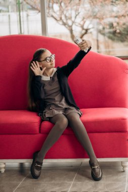 Parlak kırmızı koltukta oturan liseli kız gözlüklü ve üniformalı. Telefonlu selfie çekiyor. İç mekan, gençlik yaşam tarzı dijital eğitim kendini ifade etmek için tasarlanmış.