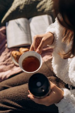 Sonbahar pikniğinde elinde çay fincanı ve termal kupa tutan tüylü beyaz kazak giyen kız. Yanında kitap ve kruvasanlarla sıcak, yumuşak dokular, rahat atmosfer.