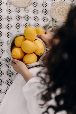 Bir kadın, karmaşık bir örgü örgü piknik battaniyesine yerleştirilmiş canlı sarı limonlarla dolu bir sepeti nazikçe tutuyor. Sahne kırsal ve rahat bir atmosfer yayıyor, doğal güzelliği ve yavaşlığı vurguluyor.