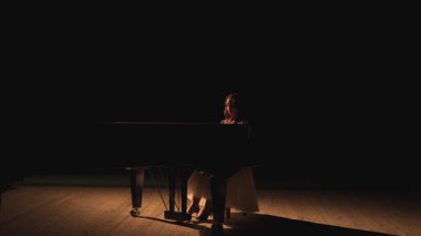 Dalgalı beyaz elbiseli zarif bir piyanist sahne ışığı altında büyüleyici ve sinematik bir an yaratıyor. Karanlığın ve yumuşak ışığın kontrastı zarafet atmosferini yükseltir.