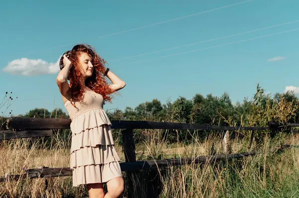 Açık renk elbiseli, kıvırcık saçlı özgür ruhlu kadın kafasında şapka, uzun çitlerin yanında duruyor. Güneşli havalarda, doğal atmosfer, kırsal enerji ve yavaş kadınsı yaz günü.