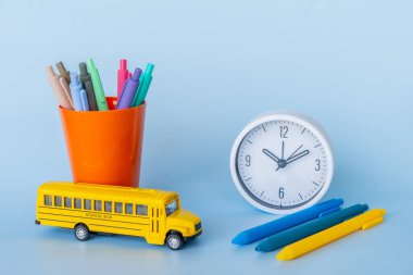 Oyuncak Otobüs ve Saatli Renkli Okul Malzemeleri Oyuncak okul otobüsü, keçeli kalemlerle dolu renkli bir kupa ve açık mavi arka planda yuvarlak bir saat. Eğitici için ideal