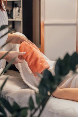 Bir spa uzmanı müşterisini nazikçe sıcak, yumuşak bir havluya sarar, rahatlama ve cilt bakımını ön plana çıkarır. Huzurlu ortam, şefkatli dokunuşu ve şımartıcı deneyimi vurgular.