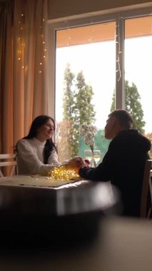 Pencere 'nin Romantik Bağlantısı genç bir çift, peri ışıkları ve çiçeklerle süslenmiş bir masanın karşısında el ele tutuşarak samimi bir anı paylaşıyor. Siluetleri yemyeşil bahçeye hafifçe parlıyor.