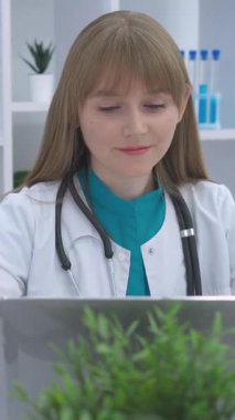 Steteskoplu beyaz önlüklü kadın bir doktor, modern bir muayenehanede dizüstü bilgisayarına odaklanmıştı. Arka planda bitkiler ve laboratuvar ekipmanları yer alıyor ve profesyonel bir atmosfer yaratılıyor..