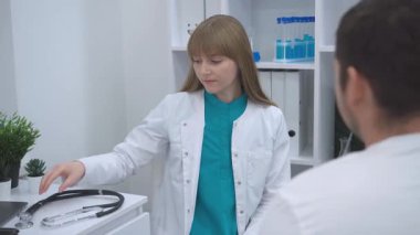 Elinde steteskop tutan beyaz önlüklü bir hastayla kadın doktor muayenehanesindeki erkek hastaya dikkatle bakıyor. arkaplanda tıbbi malzemeler ve bitkilerle raflar yer alıyor