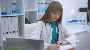 Beyaz önlüklü ve steteskoplu profesyonel kadın doktor, sağlık hizmetleri ve tıbbi belgelerin iyileştirilmesi için dijital sistemleri kullanarak parlak laboratuvar ortamındaki danışma sırasında hasta notları alıyor.
