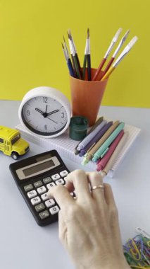 Hesap makinesi, saat, renkli kalemler ve kırtasiye malzemelerinin yer aldığı bir çalışma alanı sahnesi. Bir el hesap makinesine uzanıyor, sarı bir zemin ve enerji katıyor..