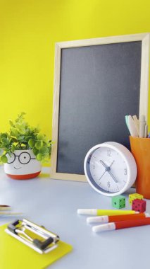 Boş bir karatahta, bir saat, renkli bir kırtasiye ve küçük bir saksı bitkisinin yer aldığı parlak ve neşeli bir çalışma alanı. Arka plan canlı sarı, enerjik bir atmosfer yaratıyor..