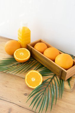 Taze bütün ve dilimlenmiş portakallar, bir şişe meyve suyu, palmiye yaprakları ve sıcak ahşap bir yüzeyde saksı bitkisi ile dengeli bir üst aşağı kompozisyon. Yerleşim, tazelik ve doğal canlılık yayıyor.