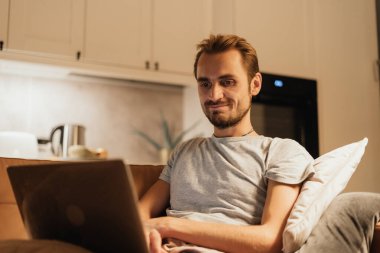 Bir adam bir dizüstü bilgisayarla rahatça oturuyor, çevrimiçi bir etkileşime dalmışken gülümsüyor. Sahne, uzak iletişimin sıcaklığını ve neşesini bir yuva ortamında yakalıyor.