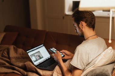 Dizüstü bilgisayarında video çağrısı ve elinde bir akıllı telefonla kanepeye sarılmış bir şekilde birden fazla iş yapan bir adam. Sıcak akşam atmosferi uzak ilişkilerin sıcaklığını ve bağlantısını vurguluyor.