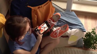 Bir bankta oturan genç bir kadın, akıllı telefon kullanıyor. O, turuncu bir çanta, mavi gömlek ve turuncu spor ayakkabılar da dahil olmak üzere günlük giyim eşyalarıyla çevrilidir. Yakınlarda küçük bir bitki var, rahat bir ortam yaratıyor.