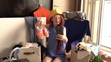 Uzun kızıl saçlı, mavi elbiseli ve sarı şapkalı bir kadın. Etrafı kıyafet ve depolarla çevrili yerde oturuyor. Elinde turuncu ayakkabılar ve telefonuyla selfie çekiyor.