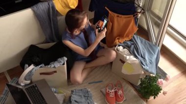 Giysiler ve çantalarla çevrili yerde oturan genç bir kadın telefonuyla bir kıyafetin fotoğrafını çekiyor. Odanın parlak, sıradan bir atmosferi var. Güneş ışığı içeri giriyor..