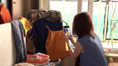 Manzaralı şık bir odada giysi eşyalarının fotoğrafını çeken bir kadın. Sahnede rengarenk elbiseler, ayakkabılar ve aksesuarlar var..