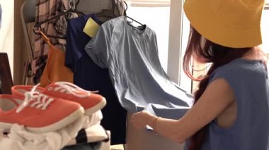 Sarı şapkalı genç bir kadın, elinde mavi gömleğiyle askıda kıyafetlerini karıştırıyor. Renkli ayakkabılar aşağıda istiflenmiş. Aydınlık ve rahat bir ortam, bir alışveriş ya da moda sahnesi izlenimi uyandırıyor.