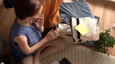 Yerde oturan bir kadın, bir kutu elbiseyi düzenliyor. Uzun saçları ve elinde bir not defteriyle gözlük takıyor. Bir bitki ve renkli giysi ürünlerinin yer aldığı ortam çok rahat..