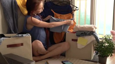 Katlanmış giysiler ve dizüstü bilgisayarla çevrili, rahat bir odada yerde oturan bir kadın. Elinde bir parça kıyafet var, düşünceli görünüyor. Arka planda bir elbise askısı ve bir bitki var.