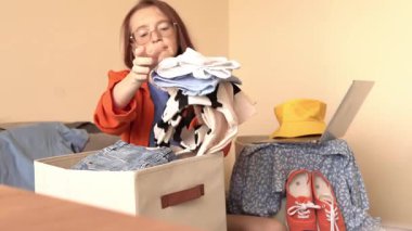 Bir kadın elinde dizüstü bilgisayarla masada otururken kutudan giysileri ayırıyor. Turuncu bir ceket giyiyor ve uzun kızıl saçları var. Yakınlarda sarı şapka ve kırmızı spor ayakkabı var..