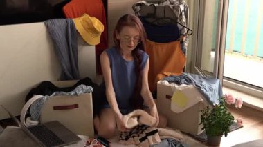 Gözlüklü genç bir kadın yerde oturuyor, çeşitli giysi ve aksesuarlarla dolu bir odada giysilerini düzenliyor. Yakınlarda bir laptop var ve bitkiler ve renkli giysiler asılı duruyor.