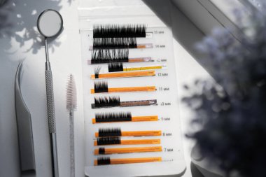 Bir tepside gösterilen kirpik uzantılarının yakın görüntüsü, 7 mm 'den 14 mm' ye kadar çeşitli uzunlukları gösteriyor. Bir kırbaç fırçası ve metal bir alet yakınlarda, bulanık çiçekli bir arkaplanda duruyor..