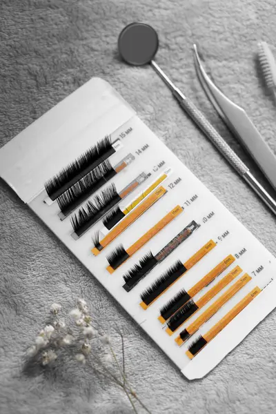 Kirpik uzantıları beyaz bir ambalajda çeşitli uzunluklarda gösterilir. Çevreleyen araçlar arasında diş aletleri ve yumuşak bir yüzeye yerleştirilmiş küçük bir buket kurutulmuş çiçek yer alıyor..