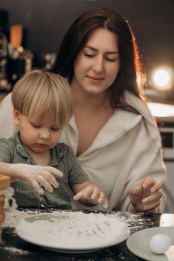 Bir anne ve bebeğinin, bir mutfak yüzeyinde unla oynadığı, sevgi ve hayal gücünü ifade ettiği, aynı zamanda çocukluk bağı ve yaratıcı neşe dolu huzurlu bir anı paylaştığı yumuşak bir sahne.